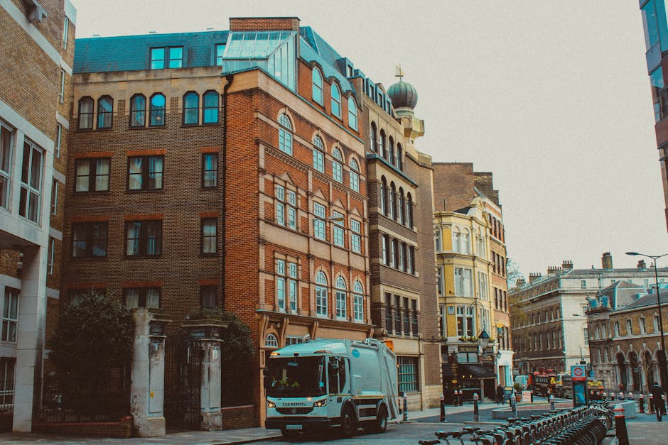 A white removal truck is parked on a city street in front of a row of multi-storey buildings with mixed brick and plaster facades, large windows, and decorative architectural details, indicating a hom