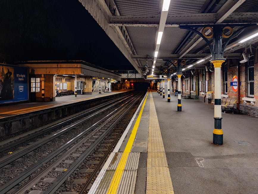 The image depicts an empty London underground train station platform at night, illuminated by artificial lighting from overhead fixtures. The platform features yellow tactile paving along the edge for